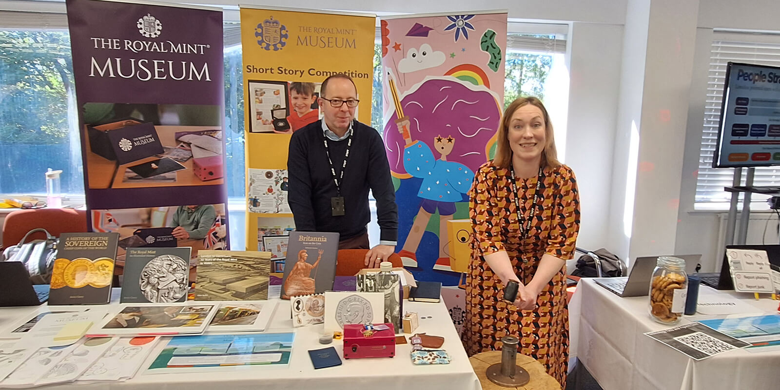 Members of the Museum Team standing behind a table full of books and pictures relating to the history of the Royal Mint, with a member of the team preparing to strike a coin using a medieval hammer and tools.