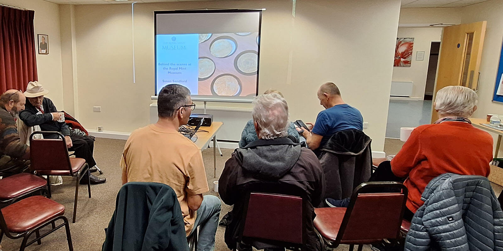 A group of people sat in chairs facing a screen with a presentation which reads behind the scenes at the royal mint museum