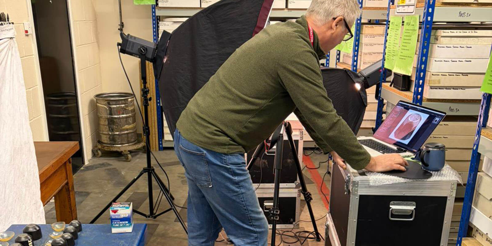 A man in a museum store room with shelves around him looking at photographs on a laptop with camera equipment behind him