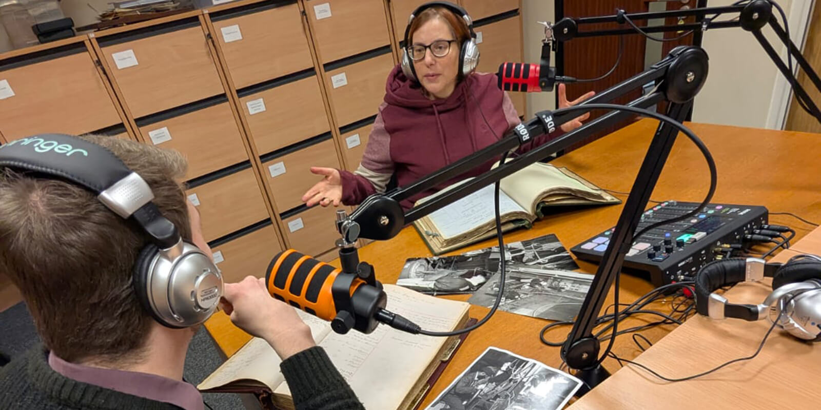 Image of two people from the Museum podcast team wearing headphones and speaking into microphones in the Museum Library with books and photographs laid out in front of them