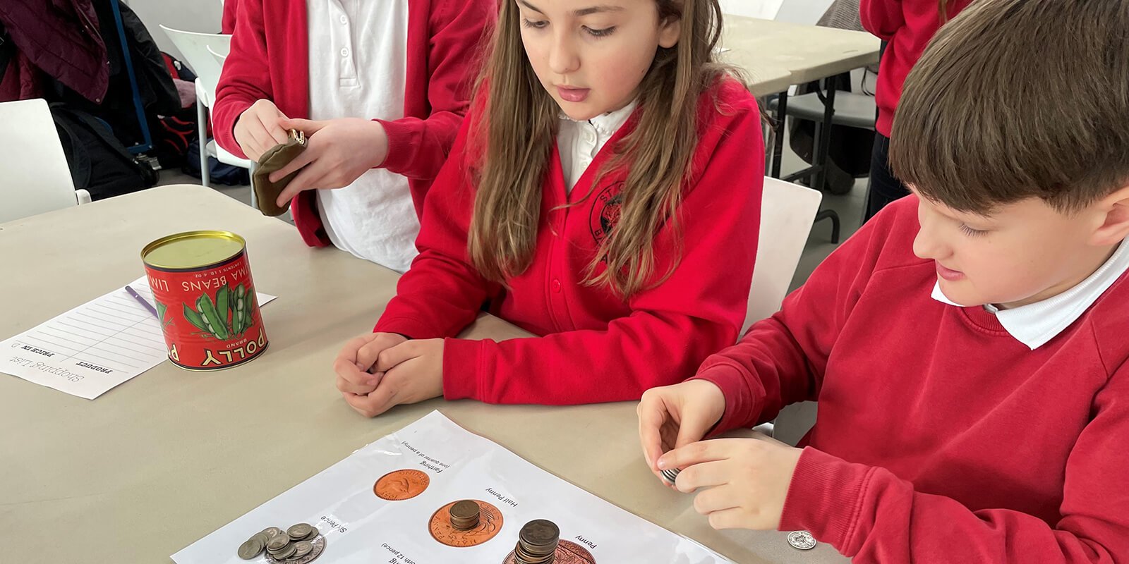 School children sit at a desk with piles of old pre-decimal coins stacked in front of them