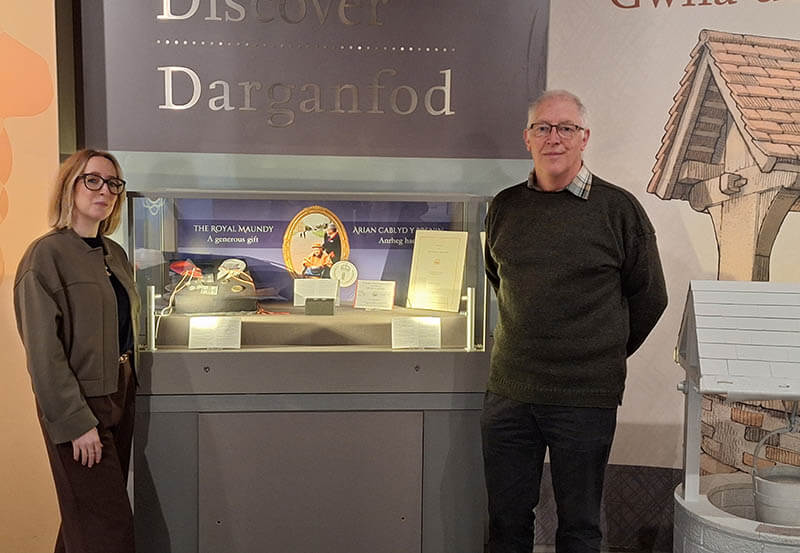 Two people standing in front of an exhibition case full of objects relating the Royal Maundy Service, each with a microphone in their hand