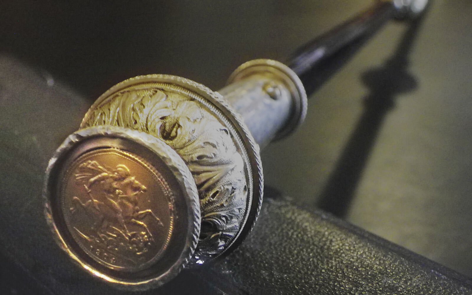 An image of a long black staff with silver details laid on a table. A gold sovereign coin is embedded in the end