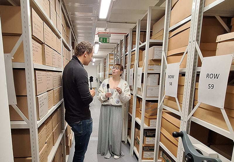 Two people standing in a storage room full of shelves of cardboard boxes, each with a microphone in theirs hand