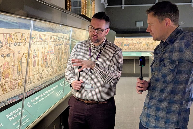 Ben Bishop and Chris Barker standing in front of the replica of the Bayeux Tapestry at Reading Museum each is hold a microphone