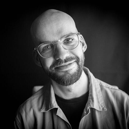 Black and white photograph of Dr Will Houstoun looking directly at the camera with his head tilted to one side